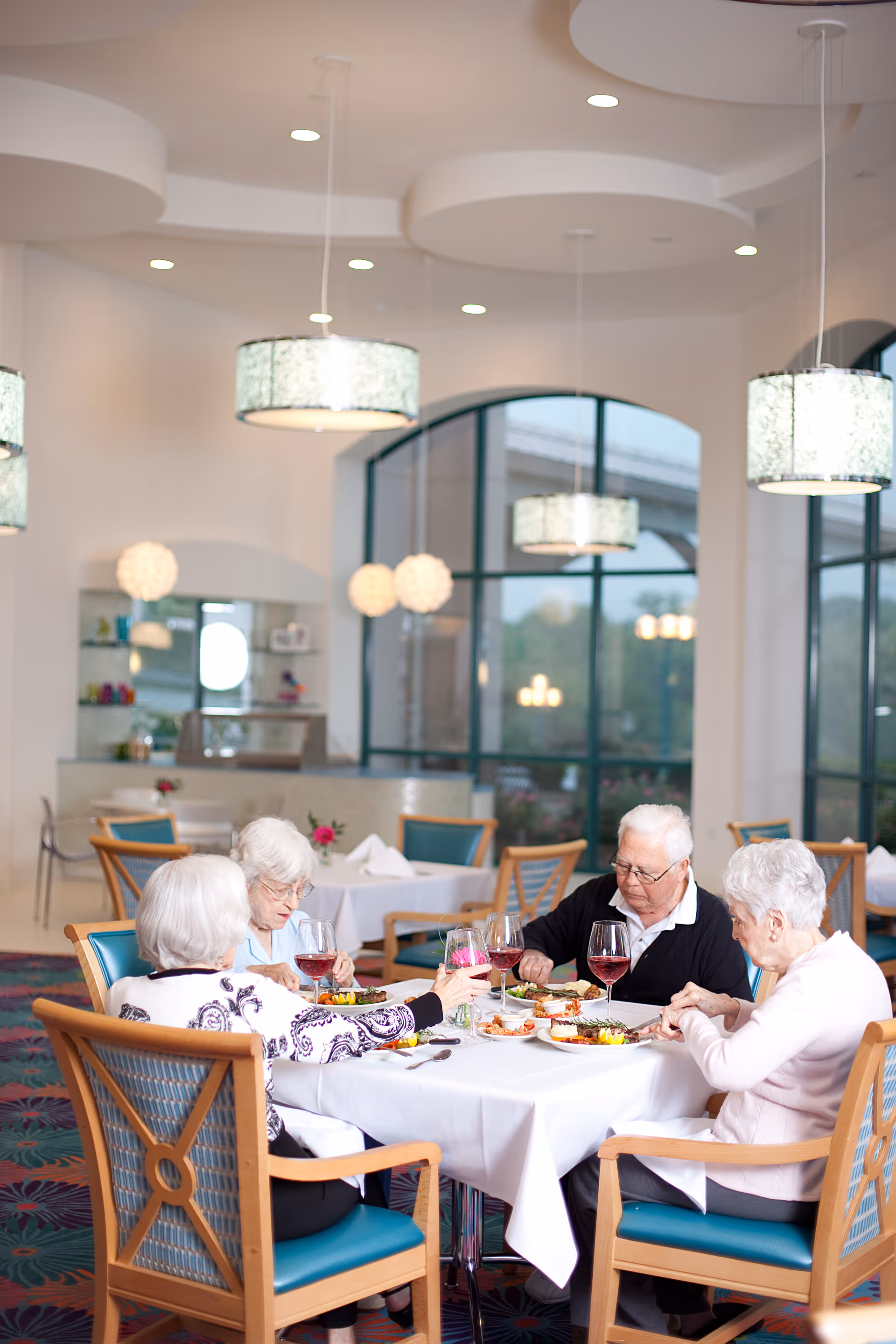 Four elderly residents seated at a table in a bright dining room enjoying a meal and wine.
