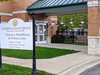 Outdoor view of St. Joseph Village of Chicago showing a sign for the Outpatient Rehabilitation & Wellness Center offering physical therapy, occupational therapy, and speech therapy. The area includes a covered walkway with hanging flower pots and benches.