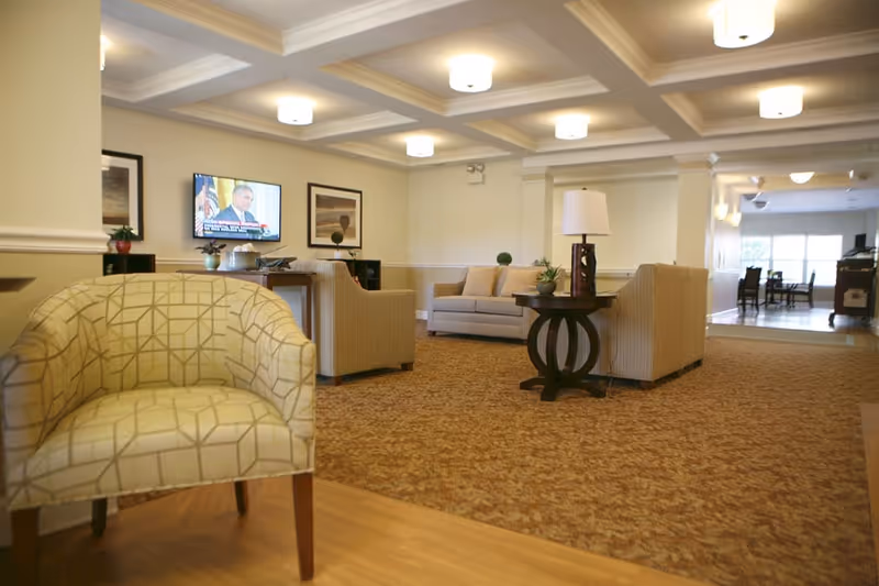 Spacious common room with upholstered chairs and sofas, a wall-mounted TV, and a side table with a lamp.