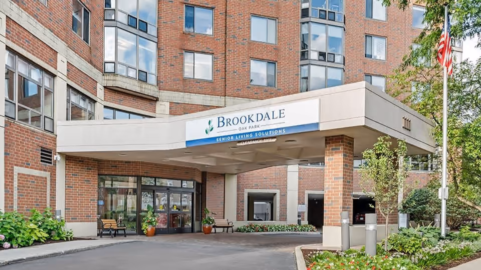Entrance of Brookdale Oak Park senior living facility showing a covered drop-off area with benches, potted plants, and a brick building facade with multiple windows. An American flag is visible on the right side near some greenery.