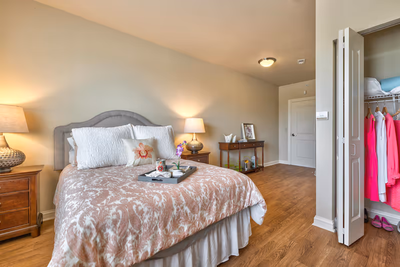A cozy bedroom with a bed covered in a patterned pink and white comforter, white pillows, and a decorative flower pillow. There are two wooden nightstands with lamps on either side of the bed. A tray with a flower, a cup, and a small plant is placed on the bed. The room has wooden flooring, a small console table with decorative items against the wall, and an open closet with clothes hanging inside. The walls are painted beige, and the room is softly lit.