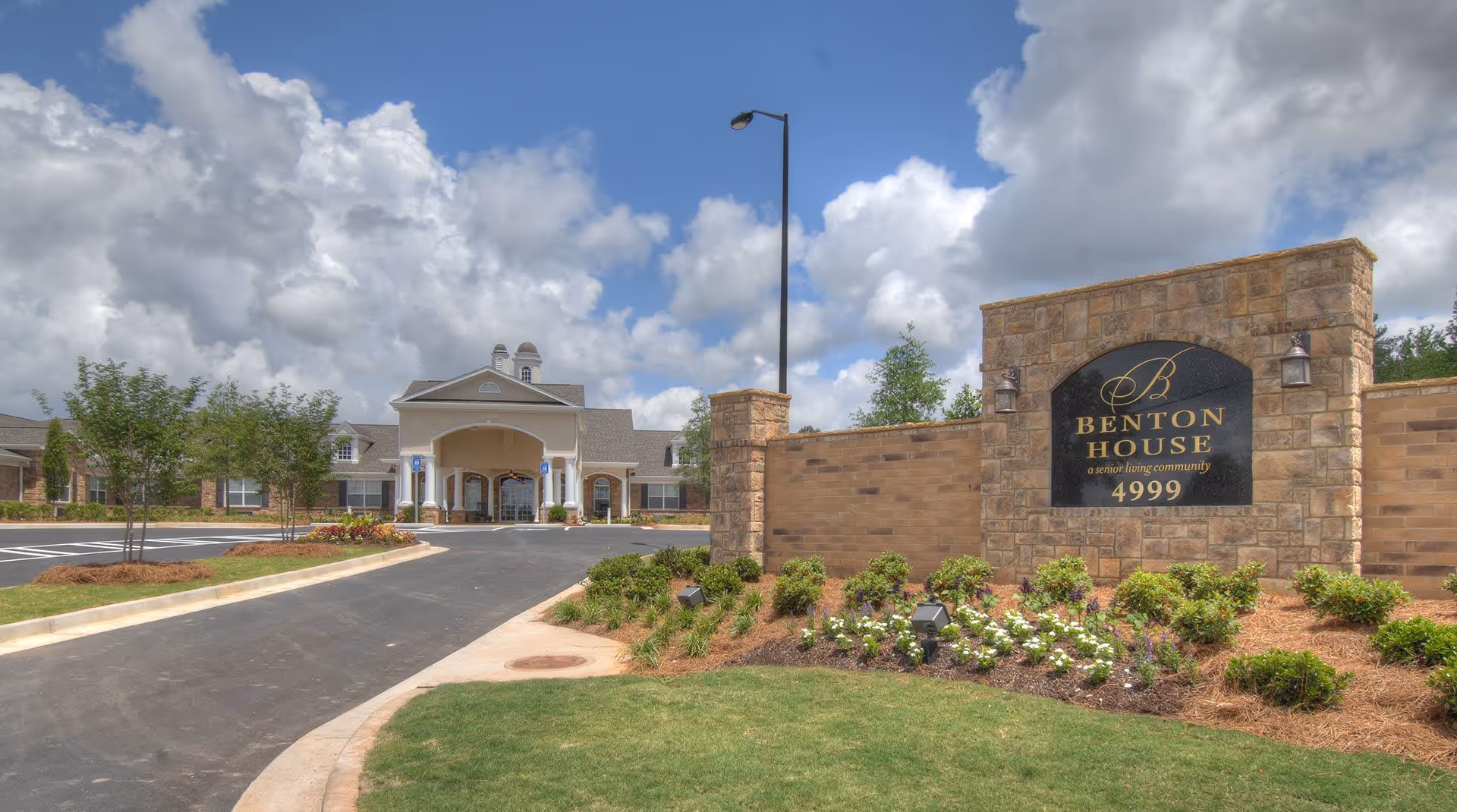 Exterior view of Benton House of Douglasville senior living community with a stone sign displaying the name and address, a driveway leading to the building entrance, landscaped greenery, and a partly cloudy sky.