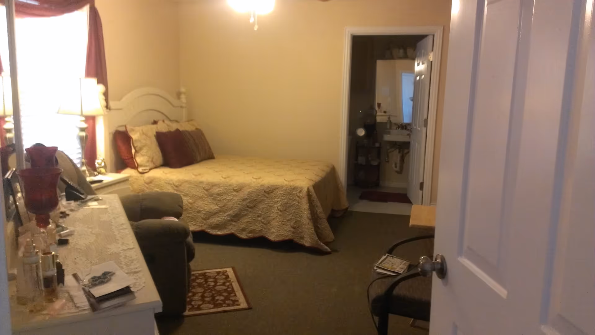 A cozy bedroom with a bed covered in a beige quilt and several pillows, a nightstand with a lamp, a dresser with various items on top, a small armchair, and an open door leading to a bathroom with a sink and mirror.
