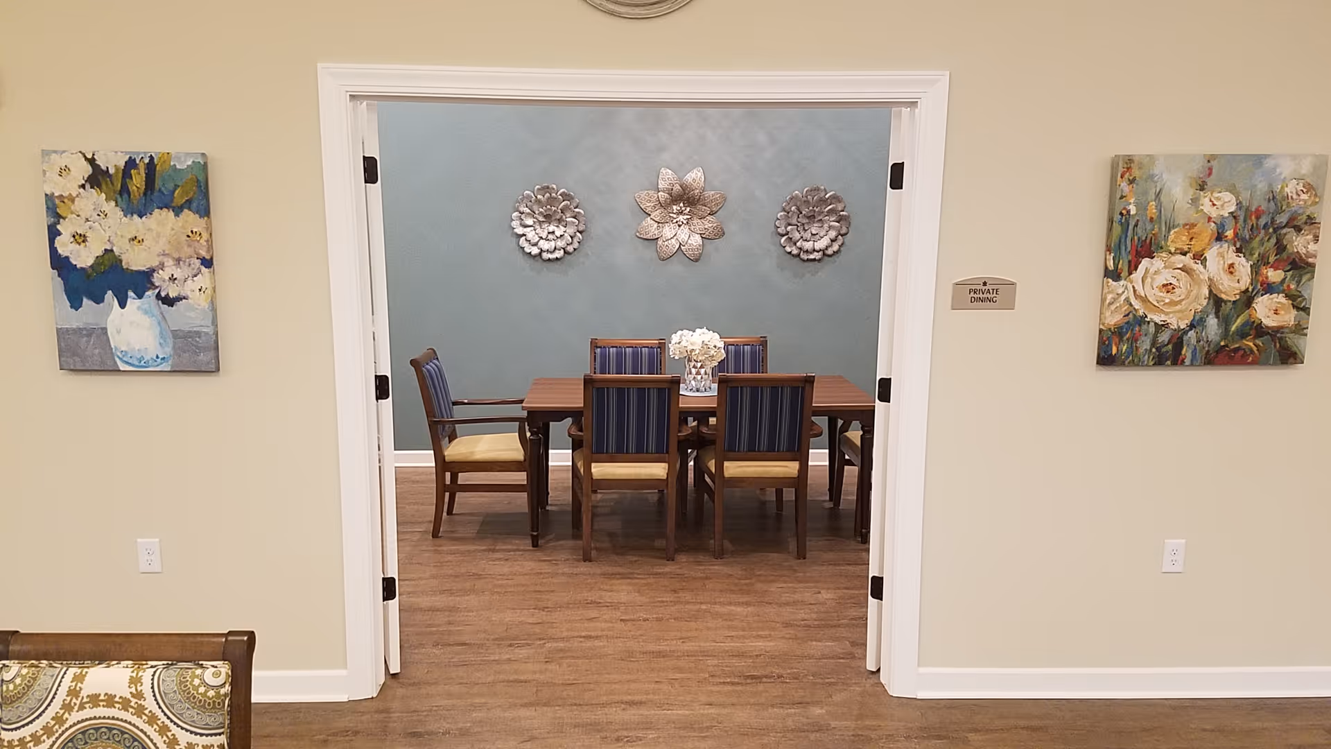 View through an open doorway into a private dining room with a wooden table and six chairs. The walls are decorated with three metallic flower sculptures and two floral paintings on either side of the doorway. The floor is wood, and a small sign next to the door reads 'Private Dining'.