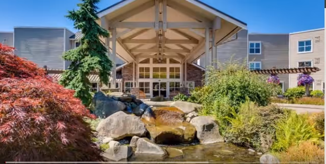 Front exterior view of a senior living facility named Crista Shores, featuring a covered entrance with large glass doors, surrounded by landscaped greenery, rocks, and a small water feature in the foreground.