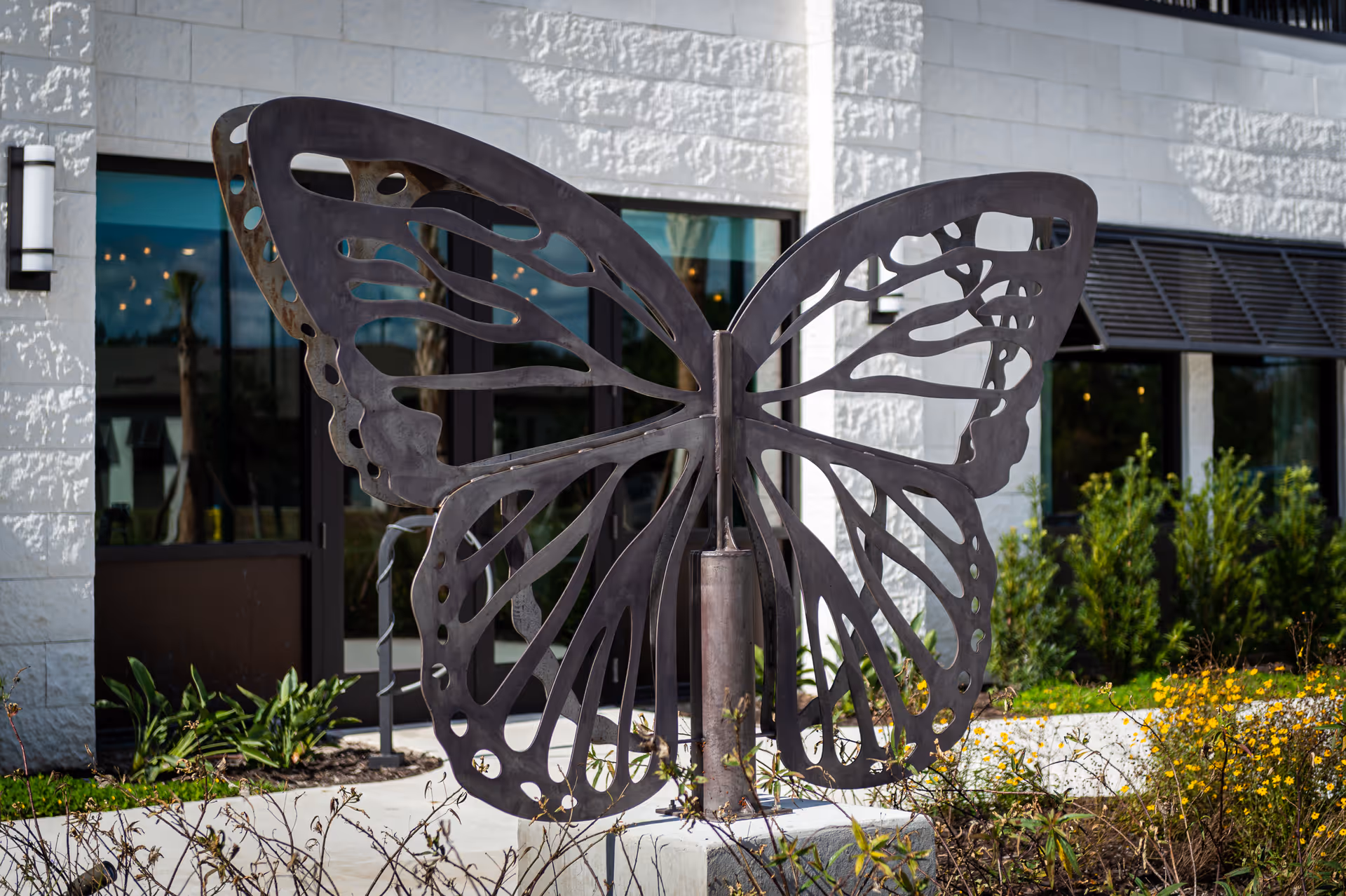 Large metal butterfly sculpture mounted on a concrete base in front of a building with white stone walls, glass doors, and windows. There are plants and yellow flowers around the sculpture.