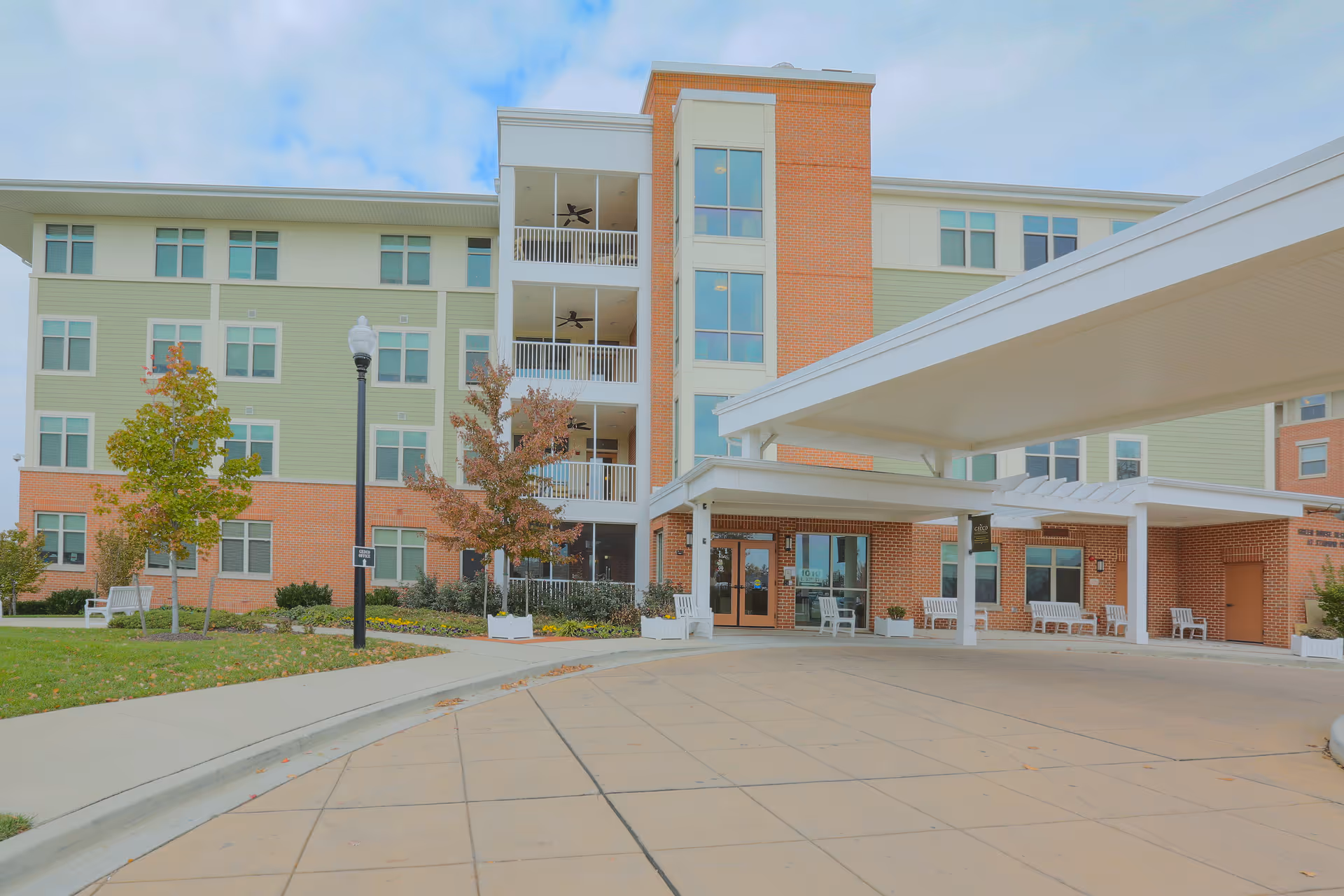 Exterior view of a multi-story nursing and rehab center building with a covered entrance driveway, several windows, a few trees, and benches near the entrance.
