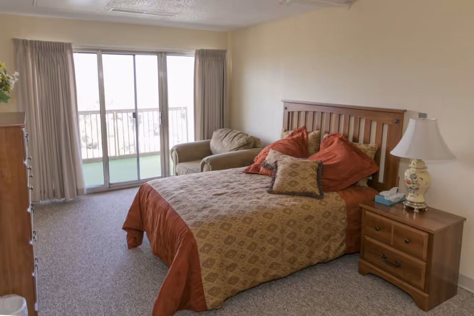 A bedroom with a wooden bed frame, a bed covered with a patterned bedspread and multiple pillows, a wooden nightstand with a decorative lamp and a box of tissues, a cushioned armchair, a wooden dresser, and large sliding glass doors with curtains leading to a balcony.