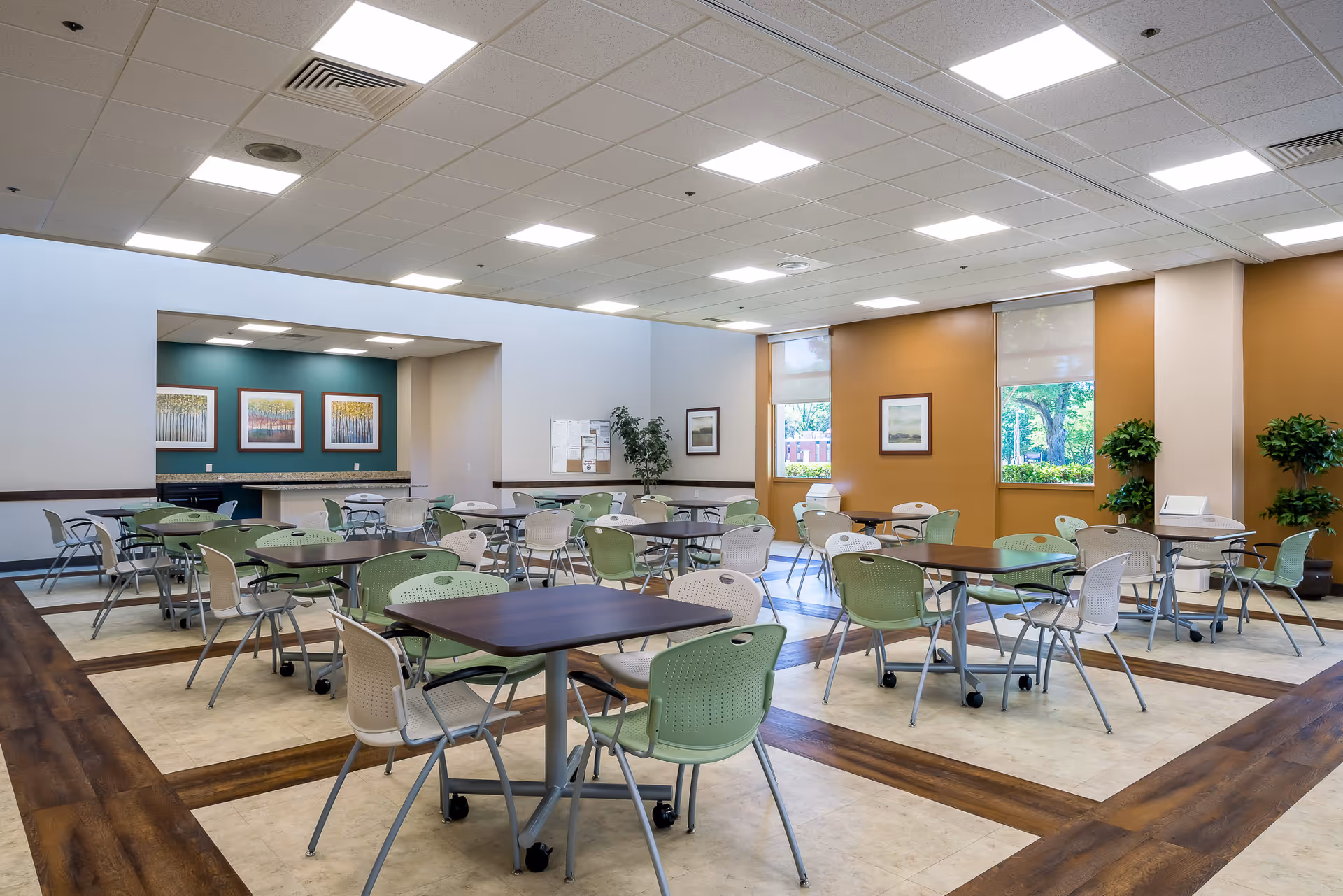Spacious bright communal dining area with multiple tables and green and white chairs, windows, and potted plants.