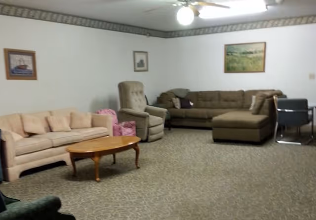 A living room with two sofas, a recliner chair, a small wooden coffee table, and a dining table with chairs. The walls are white with framed pictures hanging, and there is a ceiling fan with a light fixture.
