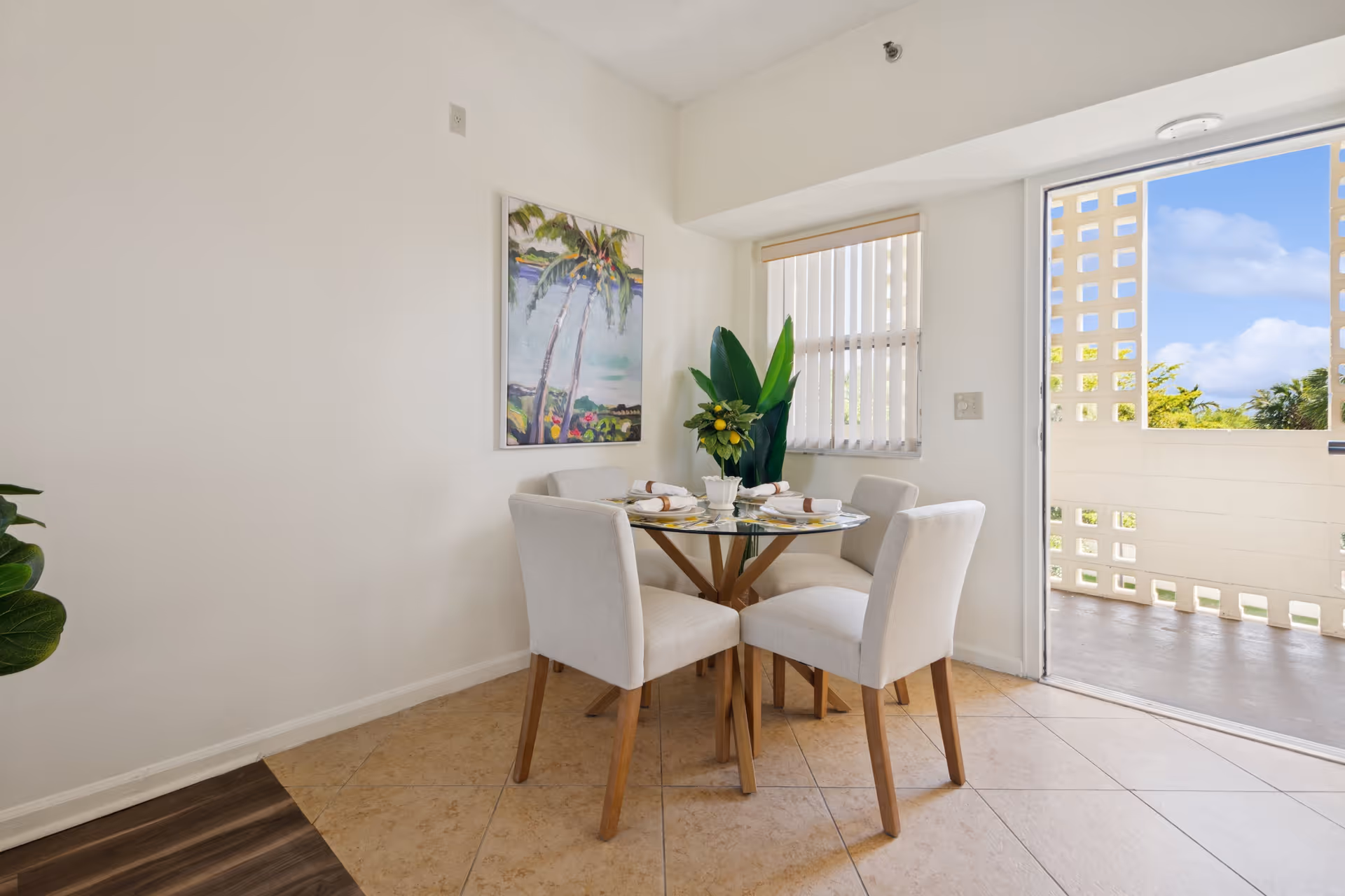 A small dining area with a round glass table and four beige upholstered chairs. The table is set with plates, napkins, and a centerpiece featuring green leaves and lemons. There is a large window with vertical blinds and a door leading to an outdoor balcony with a decorative concrete block wall. A colorful painting of palm trees hangs on the white wall.