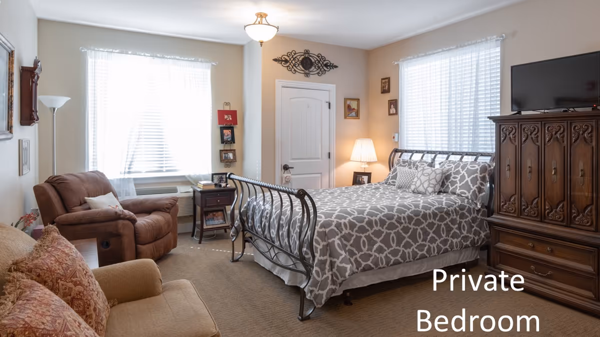 A cozy private bedroom with a metal frame bed covered in a gray and white patterned bedspread, two windows with white blinds and sheer curtains, a wooden armoire with a TV on top, a brown recliner chair, a beige armchair with patterned cushions, a small wooden side table with a lamp, and framed pictures on the walls.