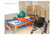 Physical therapy room with a wooden treatment table holding exercise equipment including a green exercise ball, red mat, and weights. A wheelchair is positioned nearby, and a set of stairs with handrails is visible in the background.