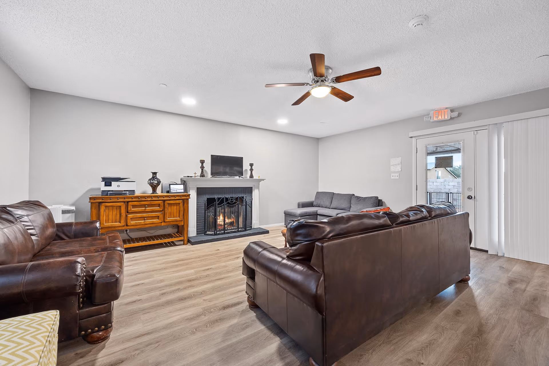 A cozy living room with a brown leather sofa and armchair, a gray sectional sofa, a wooden sideboard with a printer and decorative items, a fireplace with a fire burning, a ceiling fan with wooden blades, and a door with vertical blinds leading outside.