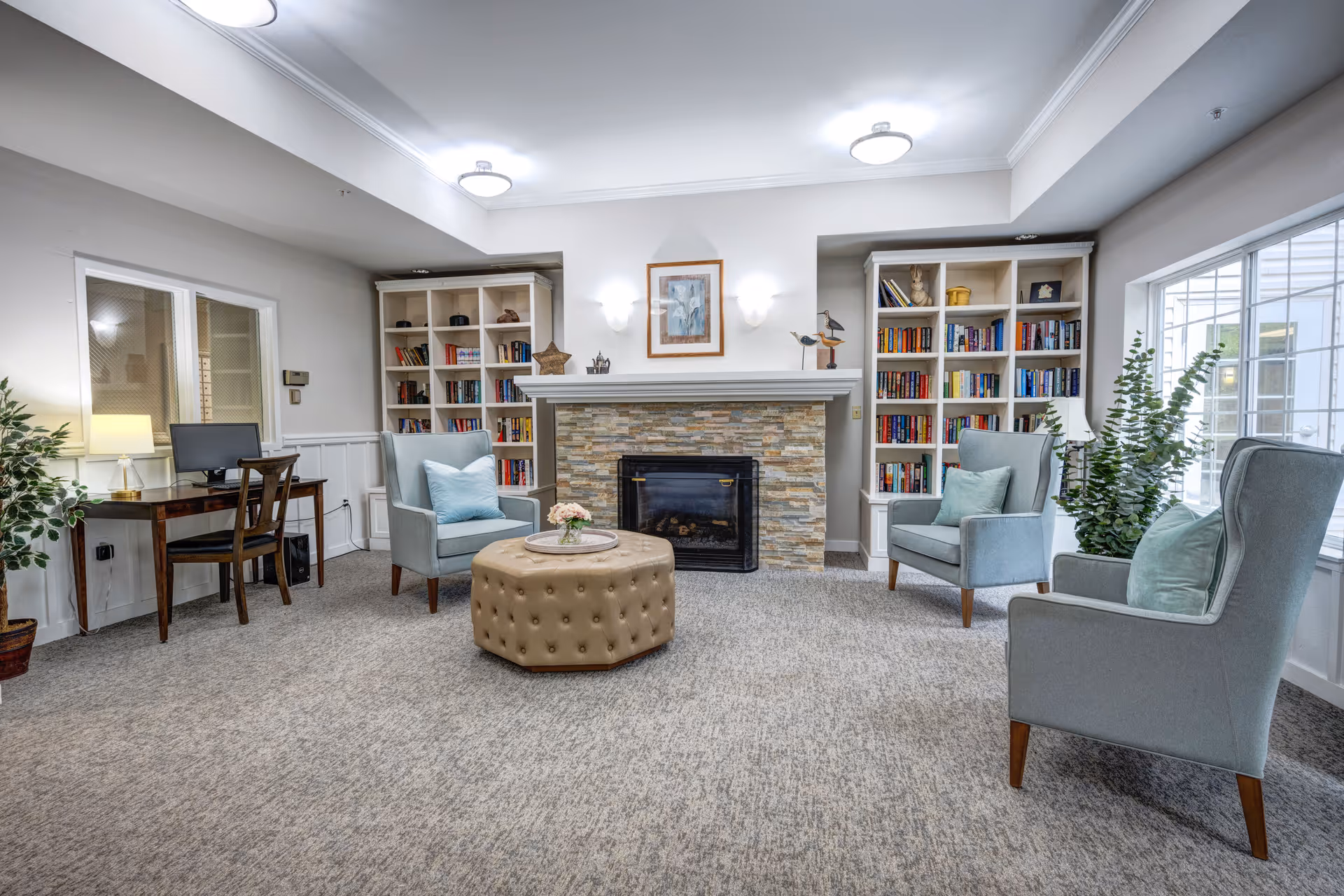 A cozy living room area with a stone fireplace centered between two white bookshelves filled with books and decorative items. There are three light blue armchairs with cushions arranged around a round tufted ottoman with a small flower arrangement on top. A wooden desk with a computer and chair is positioned against the left wall, next to a potted plant. Large windows on the right side allow natural light to fill the room.