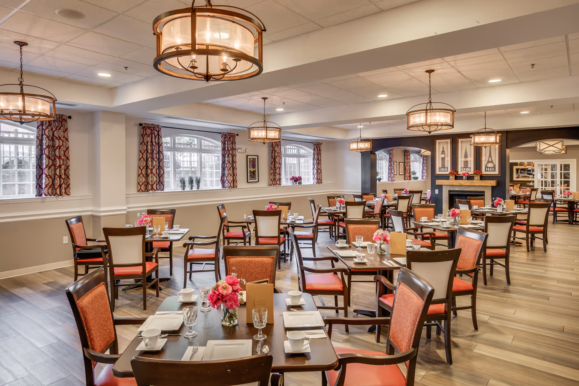 A spacious and well-lit dining room with multiple tables set for meals. Each table has chairs with red cushions, white plates, cups, glasses, and pink floral centerpieces. The room features large windows with patterned curtains, wooden flooring, and several hanging light fixtures. There is a fireplace with framed artwork above it in the background.