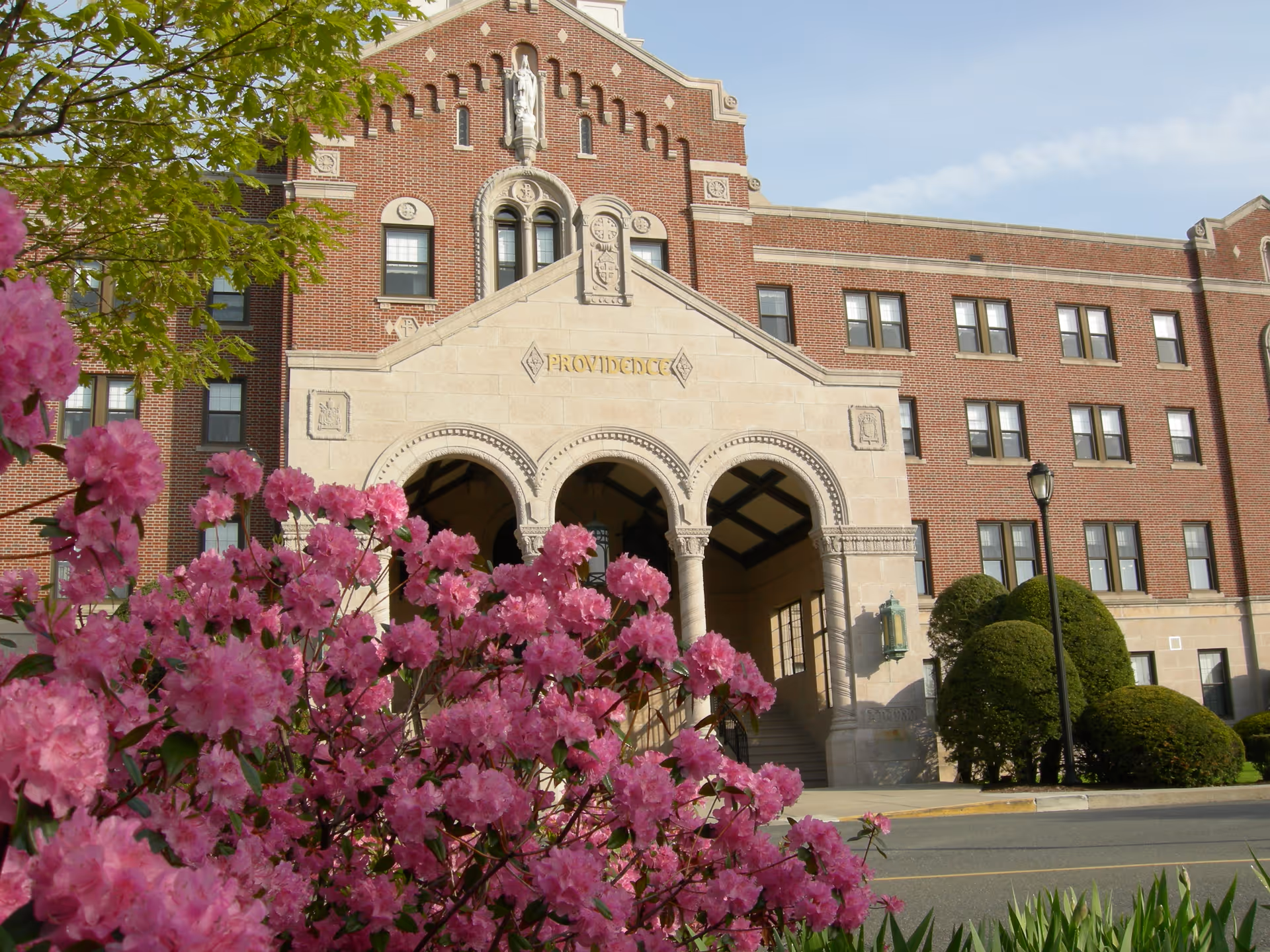 Front exterior view of a large brick building with arched stone entrance and the word 'PROVIDENCE' engraved above it. Pink flowering bushes and green shrubs are in the foreground under a clear sky.