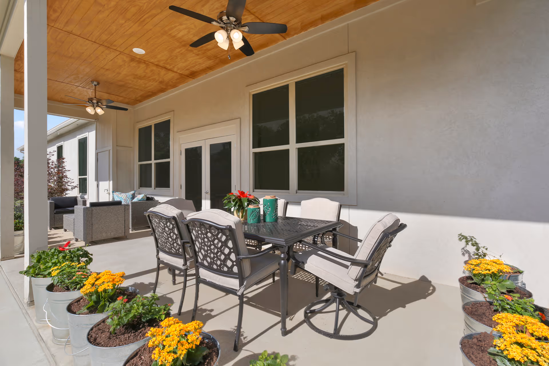 Covered outdoor patio area with a black metal table and six cushioned chairs. The patio has a wooden ceiling with two ceiling fans and is decorated with potted yellow flowers along the edge. There is additional seating with gray cushioned chairs and a small table in the background near large windows and double doors.