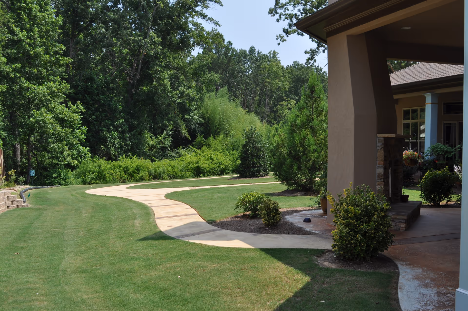 A winding concrete pathway through a well-maintained grassy area with bushes and trees, adjacent to a building with a covered patio supported by large columns.