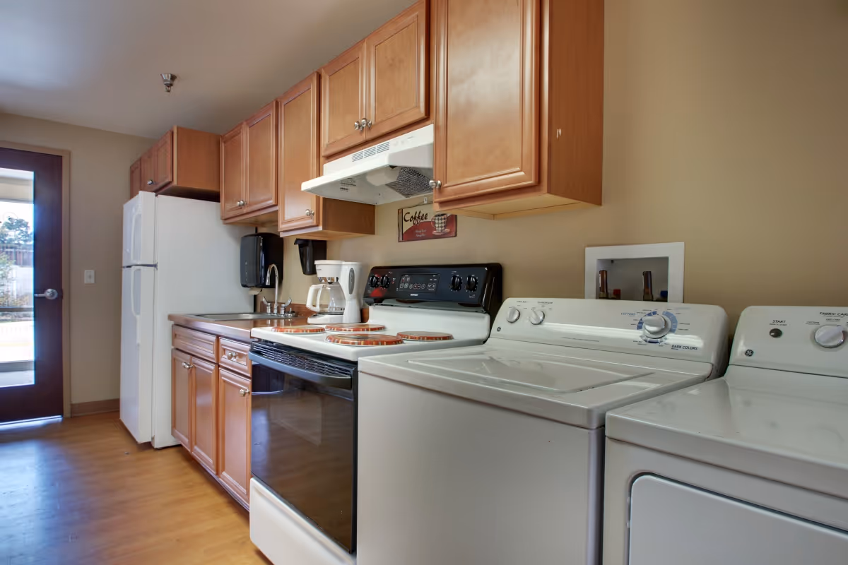 A kitchen area featuring wooden cabinets, a white refrigerator, a white stove with a coffee maker on the counter, and a white washing machine and dryer side by side. There is a door with a window on the left side letting in natural light.