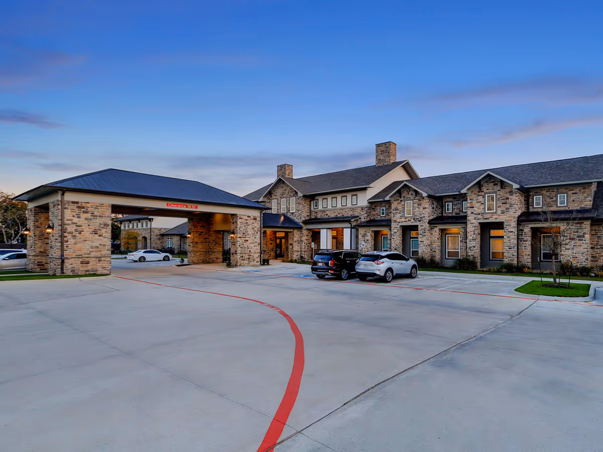 Exterior view of Pinnacle Groves facility at dusk, showing a stone building with multiple windows and a covered entrance area. Several cars are parked in the spacious parking lot in front of the building under a clear blue sky.
