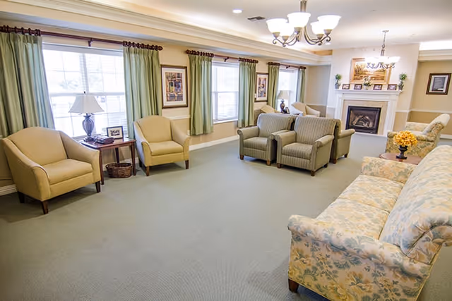 Bright communal living room with upholstered sofas and armchairs arranged around a fireplace and large windows.