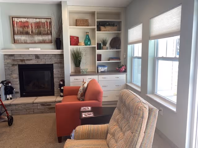 A cozy living room area featuring a stone fireplace with a framed abstract painting above it. To the right of the fireplace is a built-in white shelving unit with various decorative items including vases, books, and plants. In front of the fireplace are two armchairs, one orange and one patterned beige, with a small dark wooden side table between them. Large windows with white blinds allow natural light to fill the room.