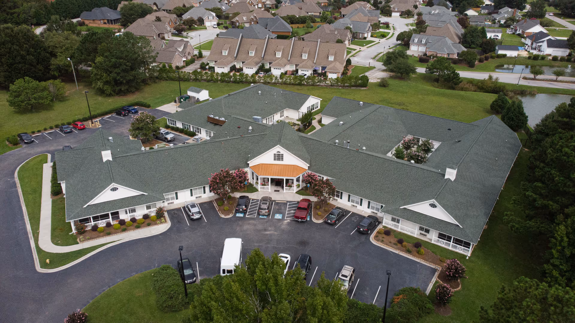 Aerial view of Pee Dee Gardens senior living facility showing a large single-story building with a green roof, surrounded by parking spaces and landscaped greenery. Residential houses and trees are visible in the background.