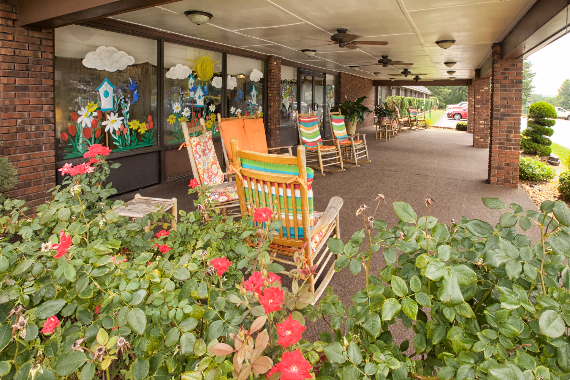 Covered outdoor patio area with several wooden rocking chairs featuring colorful cushions. The patio is adjacent to a building with large windows decorated with cheerful paintings of flowers, birdhouses, clouds, and a sun. Red flowers and green bushes are visible in the foreground, and a parking lot with cars is seen in the background.