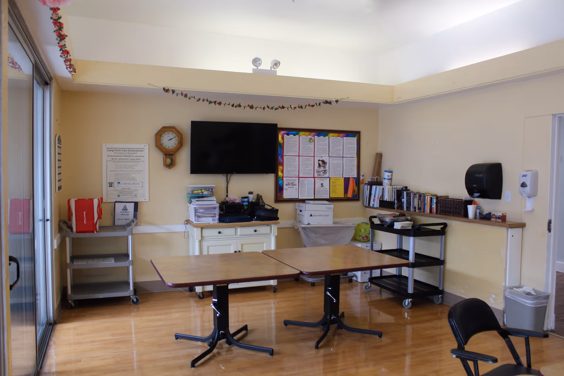 A small room with two rectangular tables pushed together in the center, a black chair in the foreground, a wall-mounted TV, a clock, a bulletin board with papers, and shelves with books and supplies. The room has wooden floors and light yellow walls with a string of decorative flowers hanging near the ceiling.