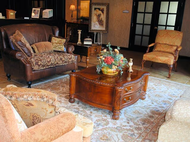 A cozy living room with a brown leather loveseat adorned with patterned cushions, two upholstered armchairs in warm tones, and a wooden coffee table with decorative carvings. The coffee table holds a floral arrangement and small decorative items. The room features a patterned area rug, a side table with a lamp, framed artwork on the wall, and double glass-paneled doors in the background.