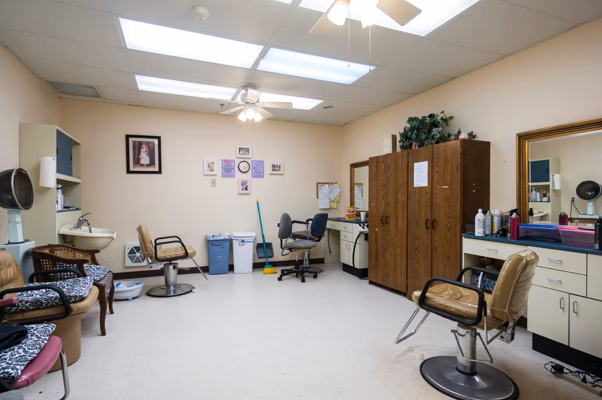 Interior view of a senior living facility's salon or grooming room with multiple salon chairs, a large mirror, a sink, cabinets, and various hair care products. The room has beige walls, a white tiled floor, and fluorescent ceiling lights with a ceiling fan. There are framed pictures and a clock on the wall, as well as a broom and trash bins in the corner.