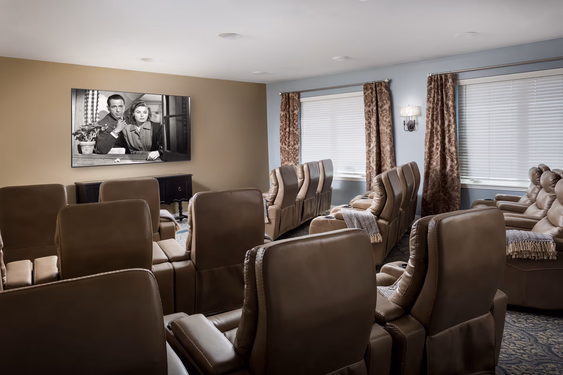 A cozy senior living facility movie room with multiple rows of brown leather recliner chairs facing a wall-mounted flat screen TV showing a black and white film. The room has beige and light blue walls, two windows with brown patterned curtains, and soft lighting from wall sconces.