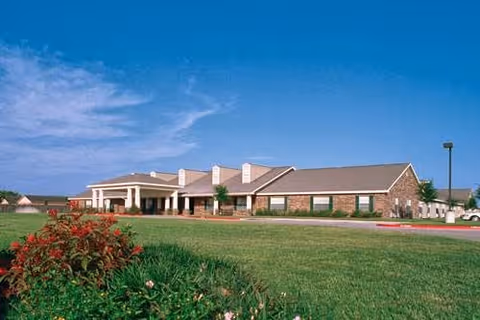 Exterior view of a single-story assisted living facility building with a covered entrance, surrounded by a large green lawn and some bushes under a clear blue sky.