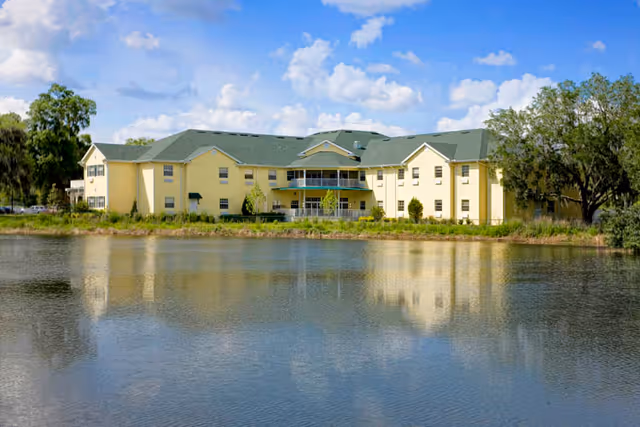Yellow three-story residential building reflected in a calm pond with trees and a blue sky.