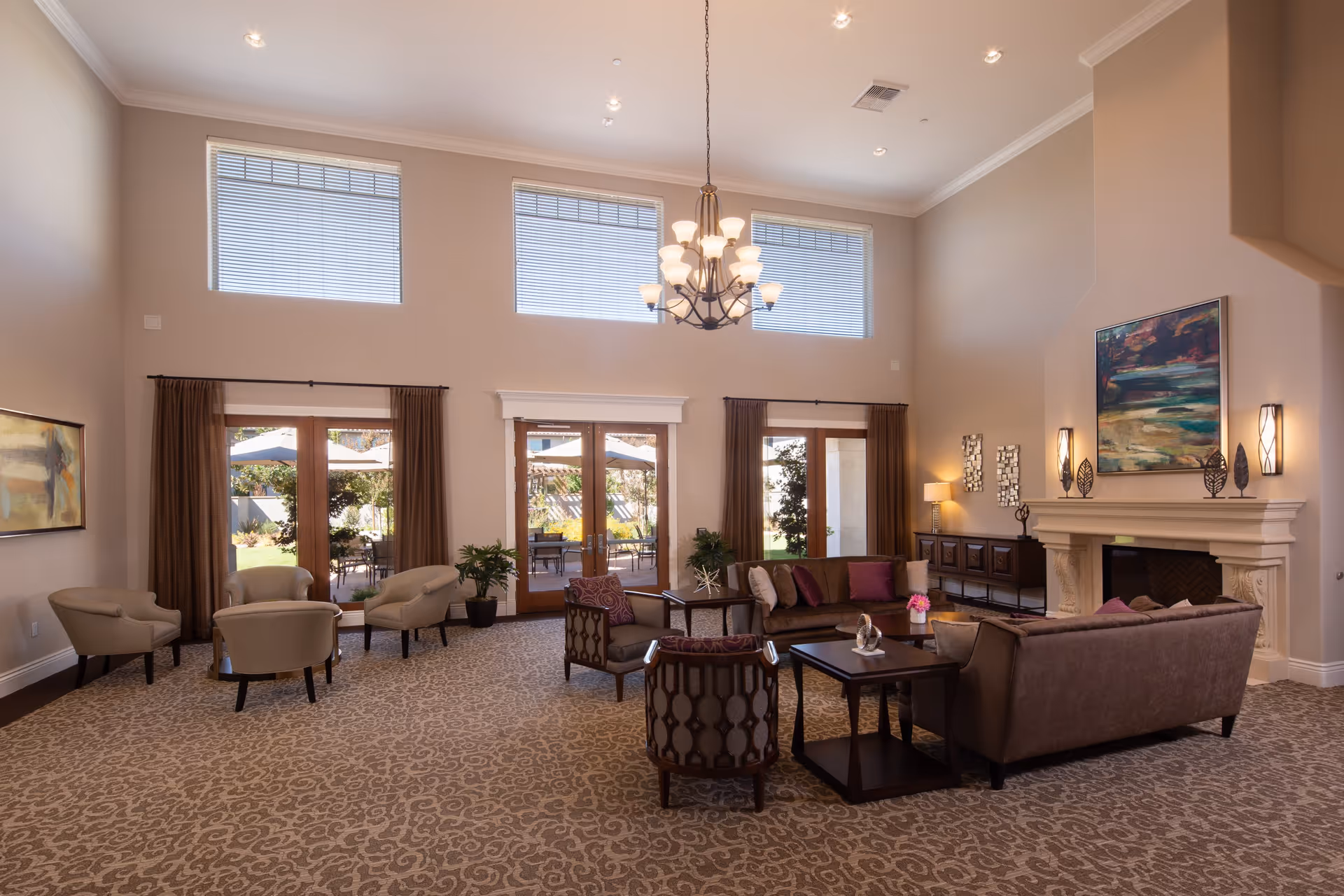 A spacious and well-lit living room with high ceilings, large windows, and glass doors leading to an outdoor patio. The room features comfortable seating including sofas and armchairs arranged around coffee tables, a fireplace with decorative items on the mantel, a chandelier hanging from the ceiling, and artwork on the walls. The carpet has a subtle patterned design, and there are plants placed near the doors.