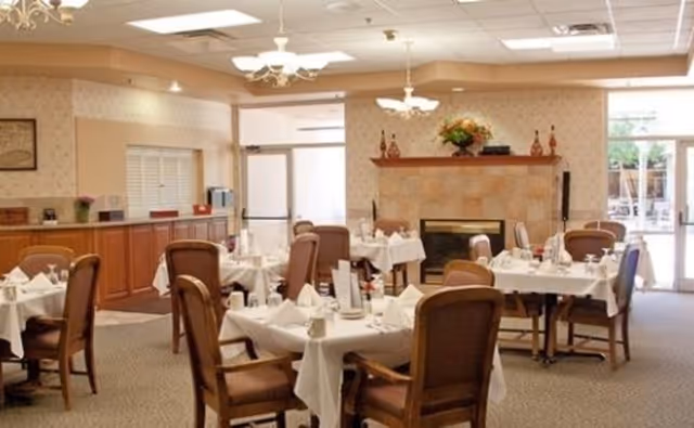 A dining room with several round tables covered with white tablecloths, each set with napkins, cups, and menus. The room features wooden chairs with cushioned seats, a fireplace with a mantel decorated with flowers and vases, and large windows letting in natural light. The walls have a light patterned wallpaper, and there is a chandelier hanging from the ceiling.