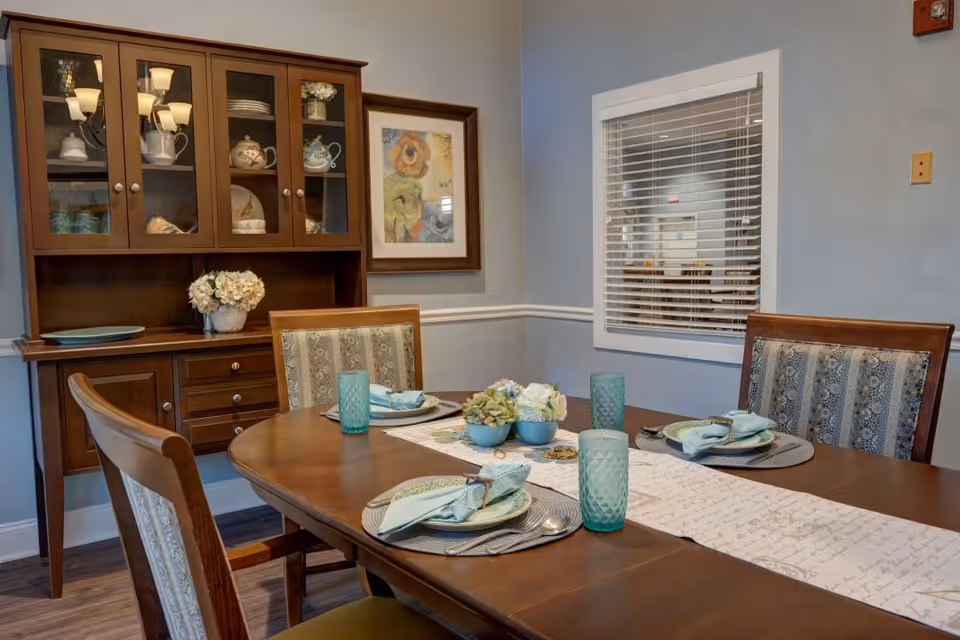 A dining room with a wooden table set for four people, featuring blue-green glasses, plates with folded napkins, and a floral centerpiece. Behind the table is a wooden cabinet with glass doors displaying dishes and a framed floral painting on the wall. A window with white blinds is also visible on the right wall.