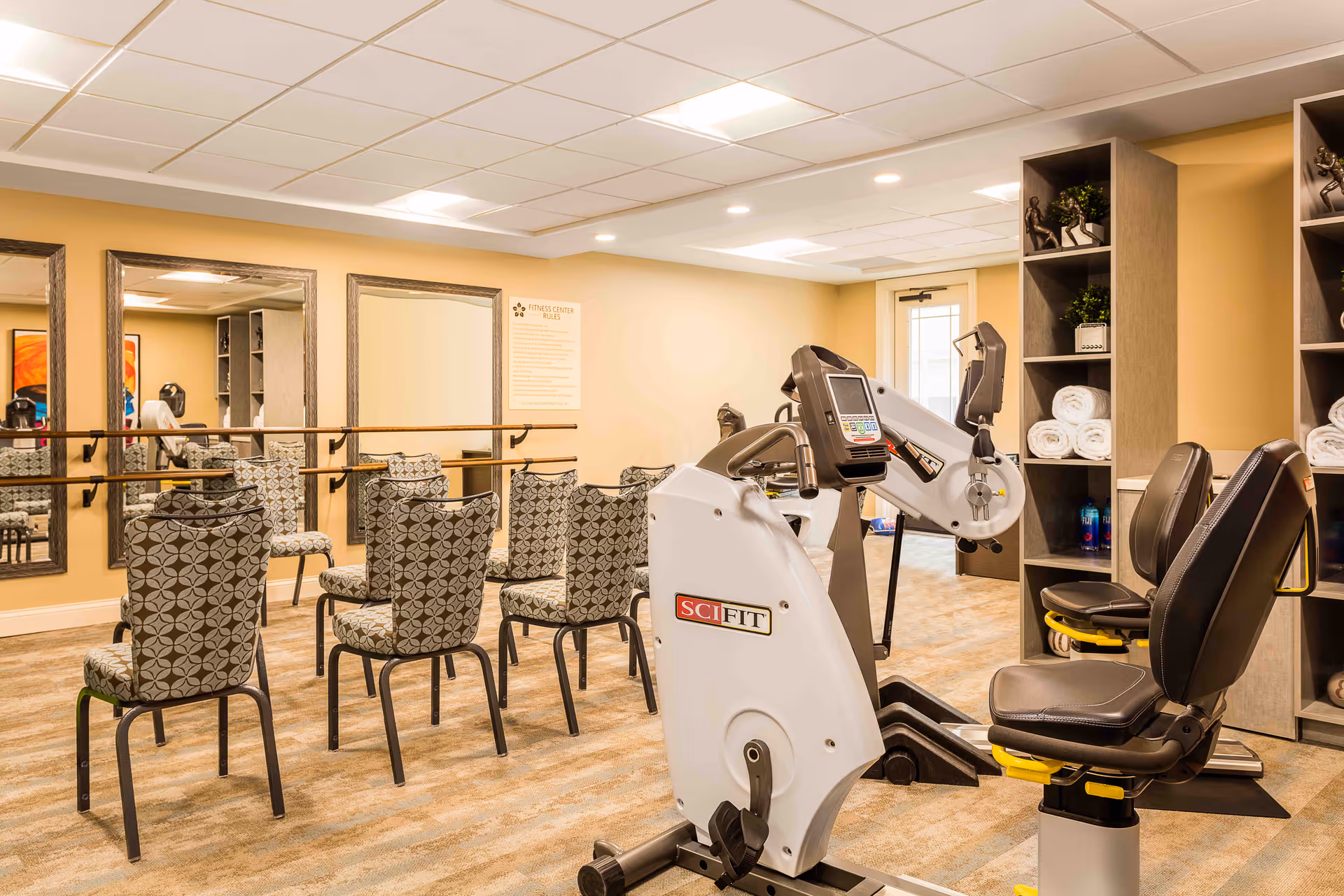 Bright assisted-living fitness room with SCIFIT exercise bikes, patterned chairs facing mirrors and a barre, and shelves stocked with towels.