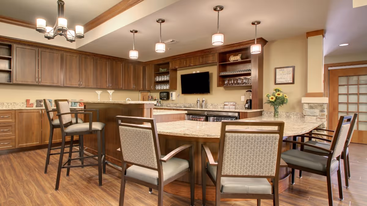 A spacious kitchen area in a senior living facility with wooden cabinets, a large granite countertop island with several chairs around it, pendant lights hanging from the ceiling, and a flat-screen TV mounted on the wall. There are shelves with glassware and decorative items, and a vase with sunflowers on the counter.