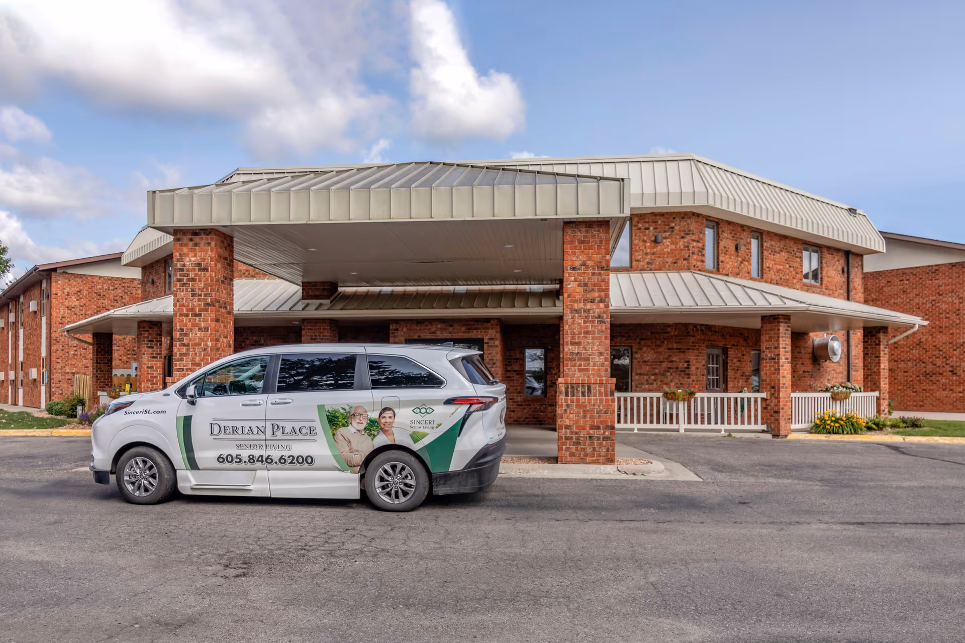 Exterior view of a senior living facility with a covered entrance supported by brick pillars. A white van with advertisements for Derian Place Senior Living is parked in front of the entrance. The building is made of red brick with a metal roof and several windows.