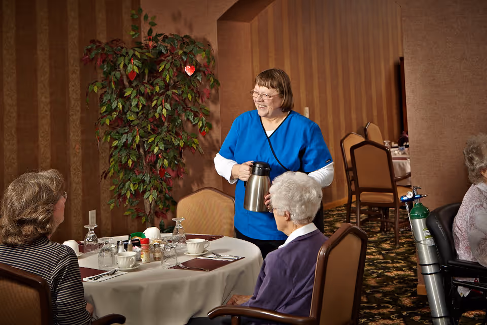 A caregiver in blue scrubs smiling and holding a coffee pot while standing next to a table where two elderly women are seated in a dining room with brown striped wallpaper and a large potted plant in the background.