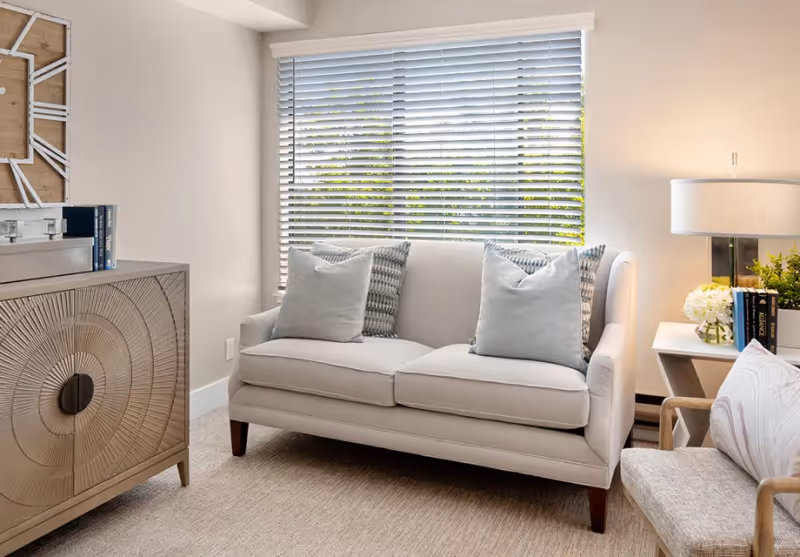 A cozy living room area featuring a light beige sofa with four decorative pillows in front of a window with horizontal blinds. To the right, there is a side table with a lamp, books, and a small flower arrangement. On the left, there is a wooden cabinet with a circular pattern design and some books on top. The room has neutral tones and soft lighting.