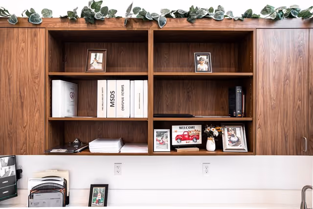 Wooden wall-mounted shelves with various items including framed photos, binders labeled MSDS and original forms, a small welcome sign with a red truck, a vase with flowers, and some stacked papers. Green leafy garland decorates the top of the shelves. Below the shelves is a white countertop with a few more framed photos and office organizers.