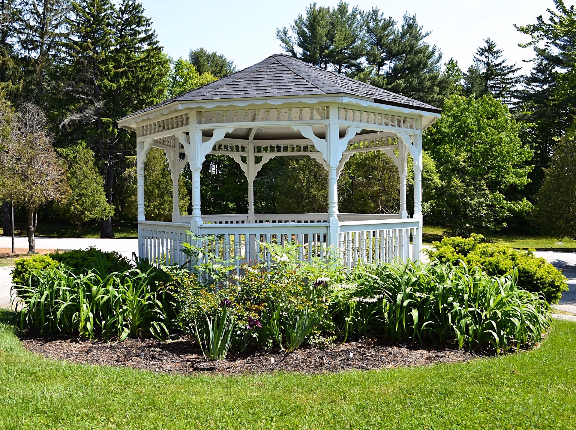 A white wooden gazebo with a shingled roof surrounded by green plants and bushes, set in a grassy area with trees in the background under a clear sky.