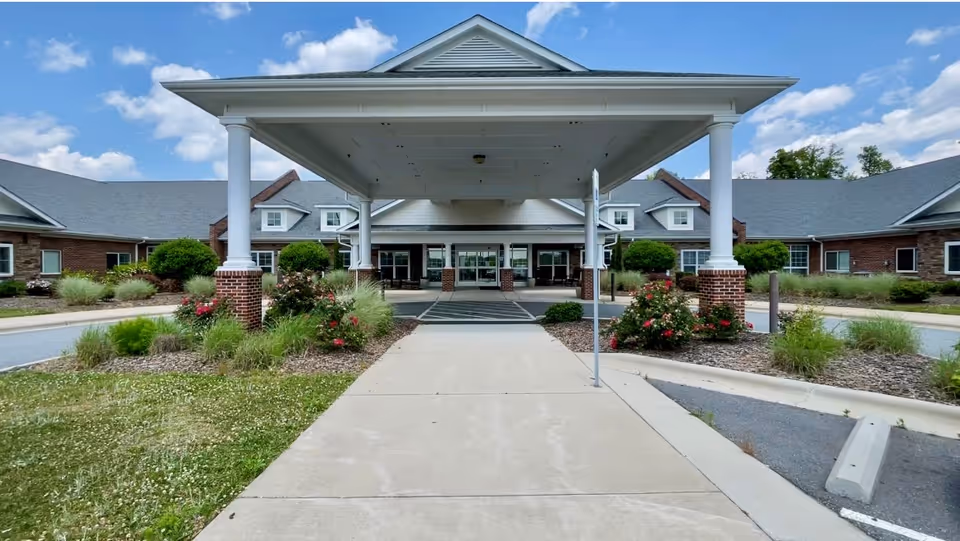 Front entrance of Summerstone Health & Rehabilitation Center featuring a covered drop-off area with white columns, landscaped bushes and flowers, and a clear blue sky with scattered clouds.