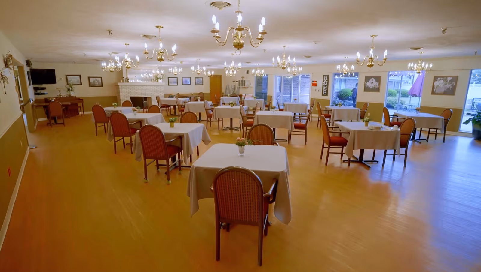 A spacious dining room with multiple tables covered in white tablecloths, each surrounded by chairs. The room features wooden flooring, several chandeliers hanging from the ceiling, and large windows letting in natural light. There are framed pictures on the walls and a fireplace in the background.