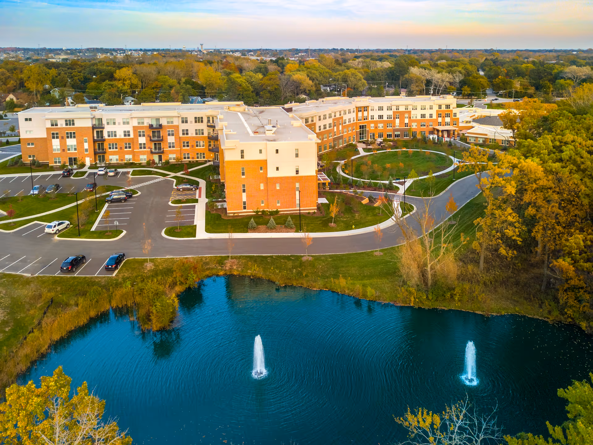 Aerial view of The Sheridan at Green Oaks senior living facility showing a large multi-story building with a parking lot, landscaped grounds, a circular driveway, and a pond with two water fountains in the foreground surrounded by trees.