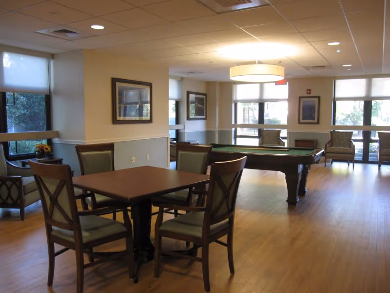 A well-lit common area with wooden flooring featuring a square table with four chairs in the foreground and a pool table in the background. The room has large windows with white blinds, framed pictures on the walls, and additional seating with armchairs near the windows.
