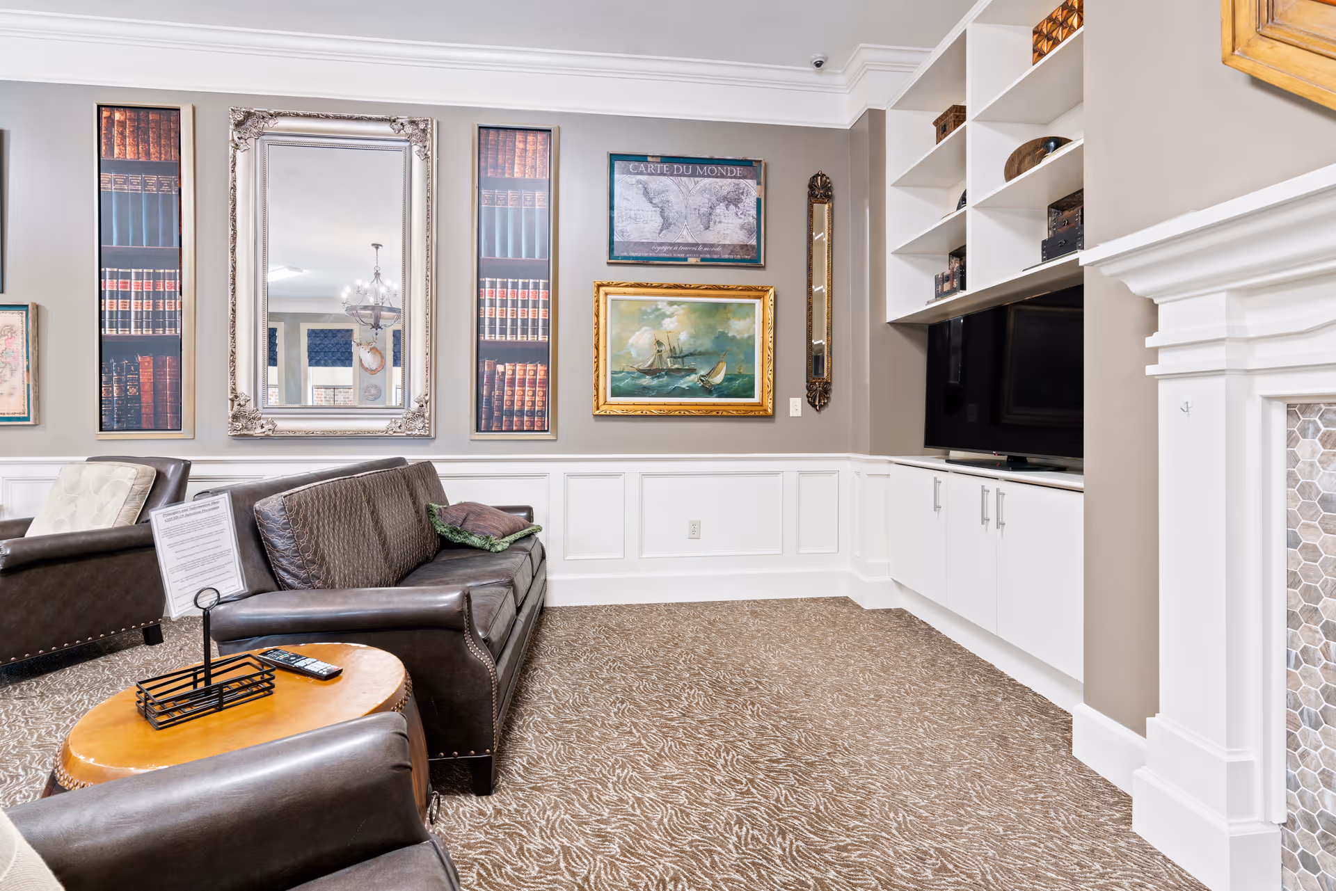 A cozy living room area with a brown leather sofa and armchair, a round wooden coffee table with a remote control and a small sign holder. The walls are decorated with framed artwork including a large mirror, paintings, and a world map. There is a built-in white entertainment center with a flat-screen TV and shelves holding decorative items. The room has beige walls, white wainscoting, and a patterned carpet. A white fireplace with a tiled surround is partially visible on the right.